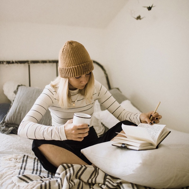 Woman sitting on her bed wearing a woollen hat and jumper, writing in a notebook while holding a cup of coffee.. Tips and Hints on Writing