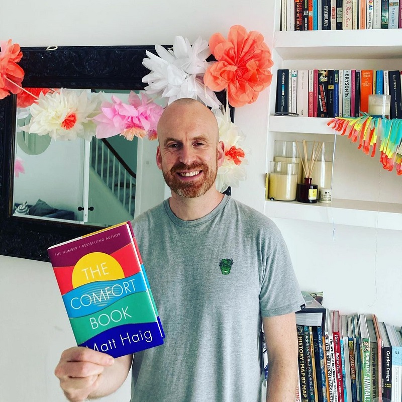 A photo* of Matt Haig holding a copy of The Comfort Book, standing in front of a bookshelf in his home.