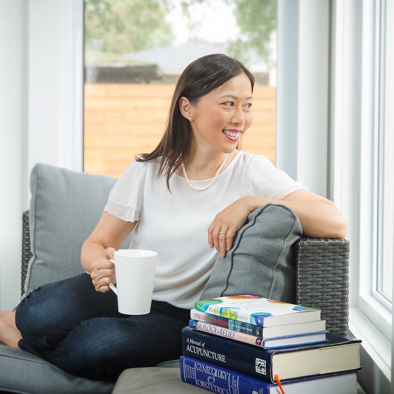 Mary Wong is sitting in a grey chair in dark blue jeans and a white t-shirt. She is holding a white mug and is looking out the window. To her right is a stack of five books.