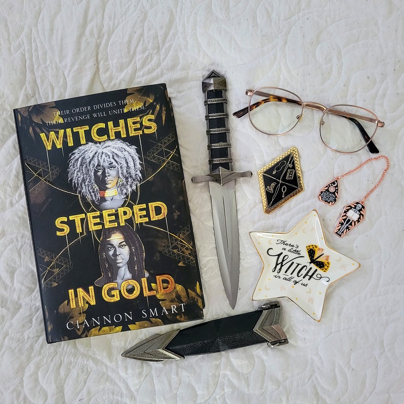 A copy of Witches Steeped in Gold with a dagger, sheath, glasses, enamel pin, chain bookmark, and jewelry dish.