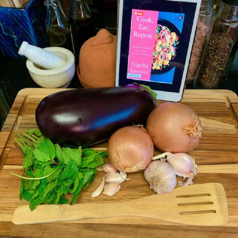 Image shows the front cover of Cook, Eat, Repeat by Nigella Lawson displayed on an iPad surrounded by cooking utensils, an eggplant, onions and head of garlic