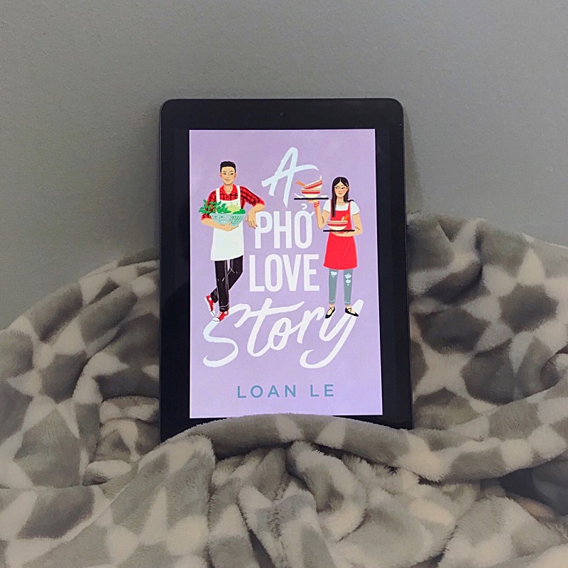 The lavender cover of A Pho Love Story is shown on an iPad screen. The iPad rests on a gray and white blanket, and leans against a blue-gray wall.