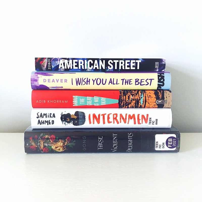 Best YA Book Club reads: The cover of These Violent Delights faces outward with its rose, gold, and black design. On the right, the spines of Internment, Darius the Great is Not Okay, I Wish You All the Best, and American Street all stand in a row. The books are on a white desk and the background is a light blue-gray.