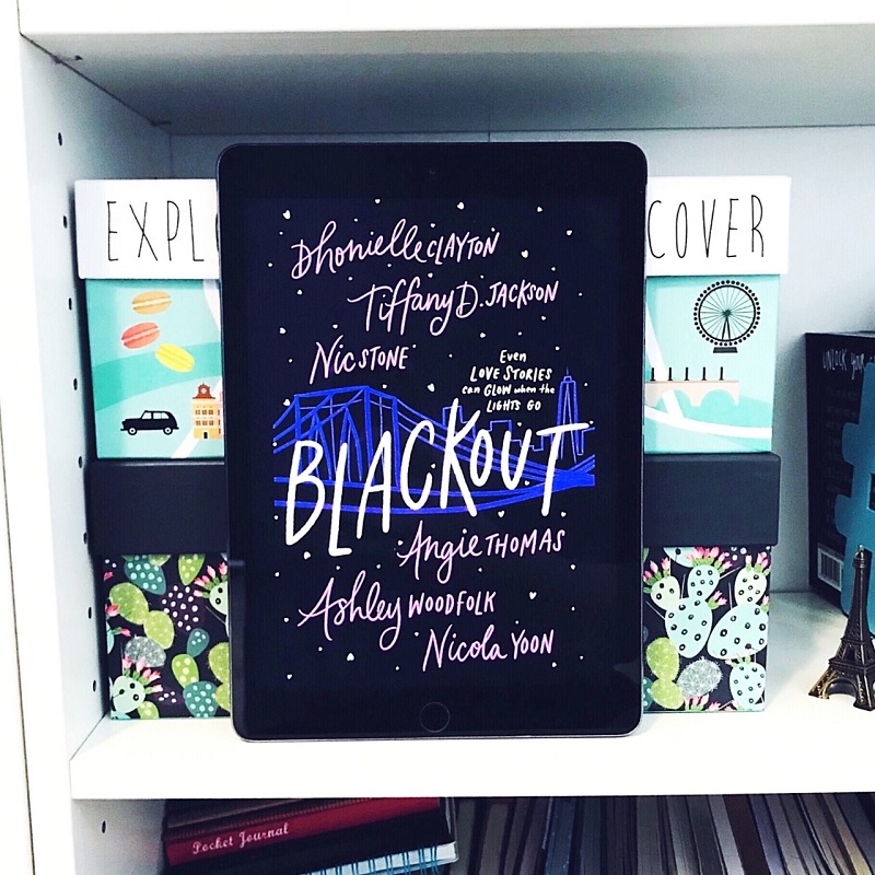 The dark cover of Blackout is shown on an iPad screen. The title is in white, and the authors names are in purple, with a blue bridge and skyline in the middle. The iPad leans against two photo boxes, which all sit on a white bookshelf.