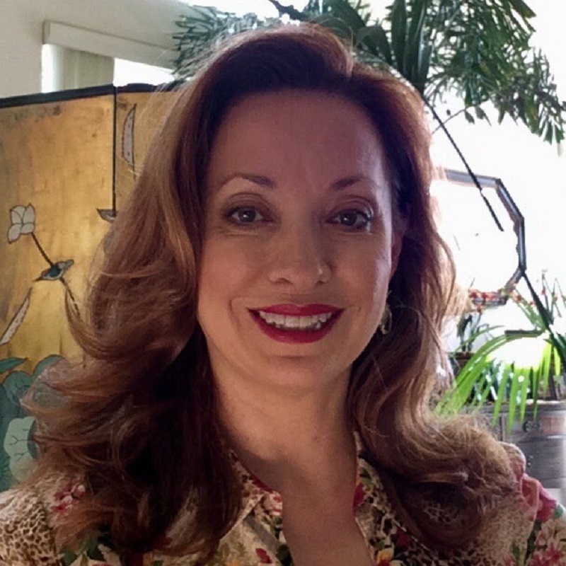 The author, Jan Moran, is centred. She has shoulder length brown hair, is wearing red lipstick and has on a cream-coloured floral shirt with leopard print. Behind her is a wooden cupboard with a floral design, tall green plants and a window.