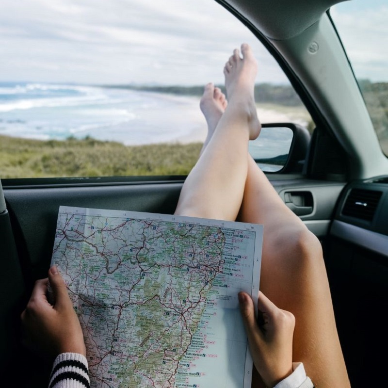 ”woman laying back in a car with crossed feet out the window and a map in her hands. Beachfront in the background.”Summer Self-Care Staycations in Ontario