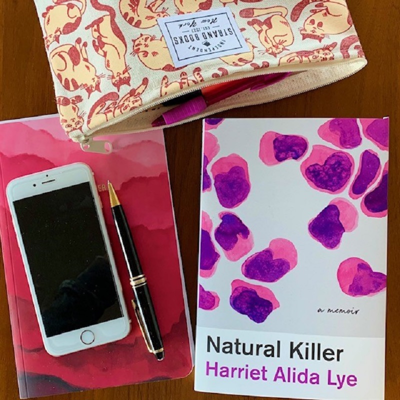 A copy of Natural Killer by Harriet Alida Lye next to a notebook, pen, pencil case and cell phone on a desk