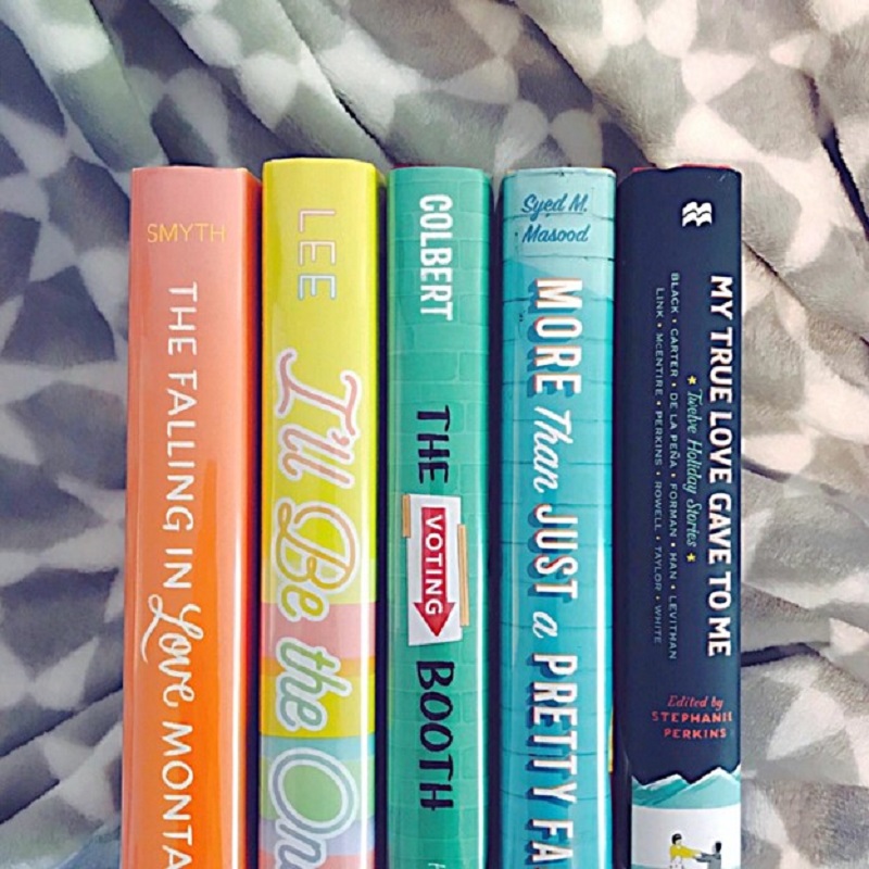Five book spines are lined up on a geometric gray and white blanket. The top two-thirds of the books are showing, and the spines are orange, yellow, green, blue, and navy, therefore forming a rainbow.
