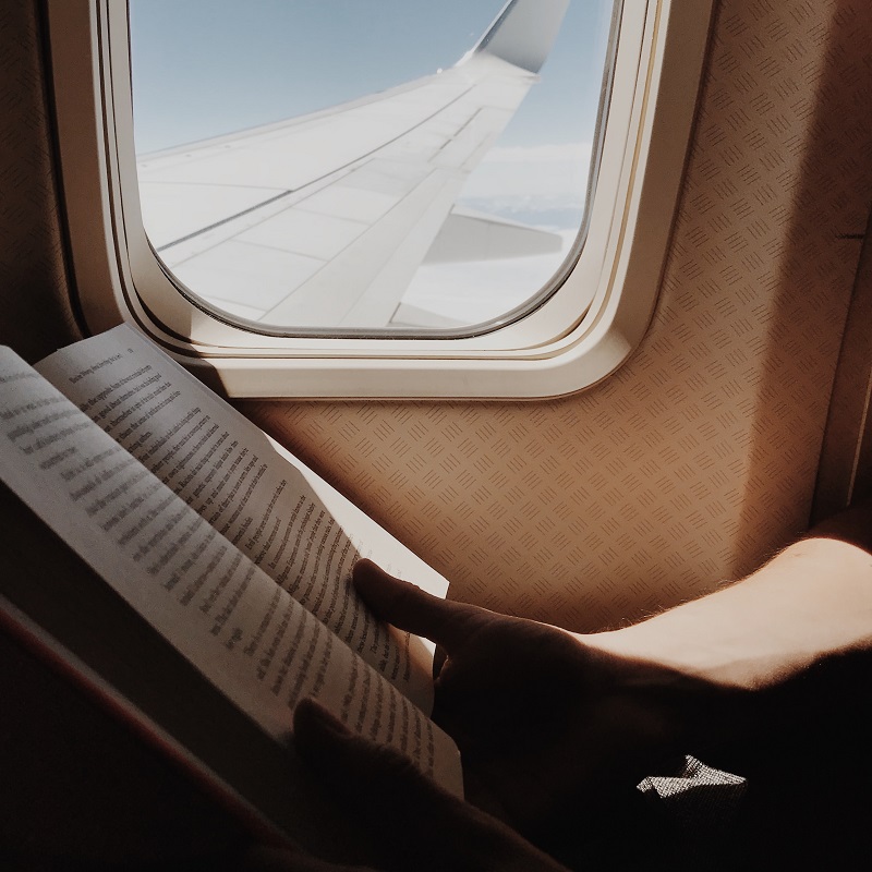 five carry-on reads to enjoy on a plane. A book held open by the window of a plane with the plane wing in the background.