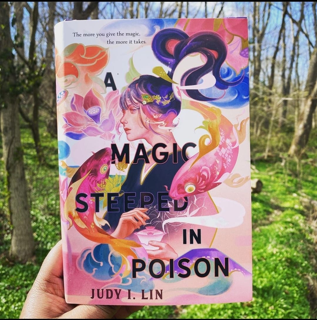 – A copy of A Magic Steeped in Poison held by a hand in the woods. The cover shows an Asian woman with purple hair in a swirling ponytail holding a teacup. The background features koi fish and lotus flowers.