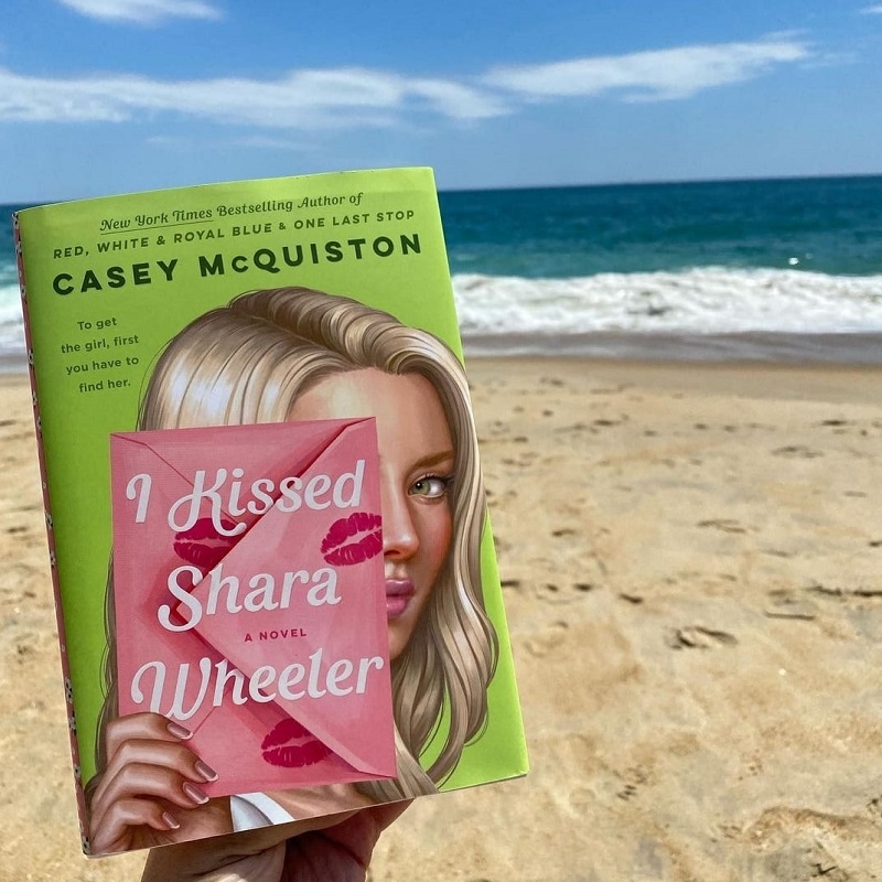 – A copy of I Kissed Shara Wheeler being held in front of a sandy beach and blue ocean below a blue sky. The cover of the book shows a blond girl with a white face. Her face is mostly covered by a pink envelope with red lipstick kisses. The title of the book is in white letters on the envelope. The background of the book cover is lime green
