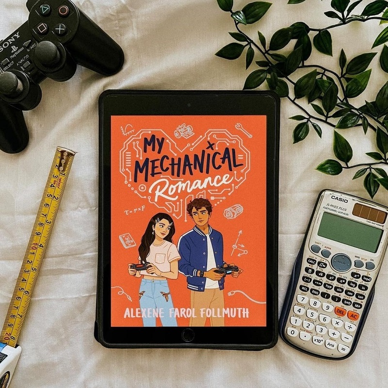 black Playstation controller, a yellow measuring tape, a green vine plant, a silver calculator, and an iPad lay on a ruffled white blanket. The iPad displays the cover of My Mechanical Romance. A Filipino-American girl in a pink t-shirt and jeans holds a controller and looks over her shoulder at a Mexican-American boy in a blue jacket and khaki pants.