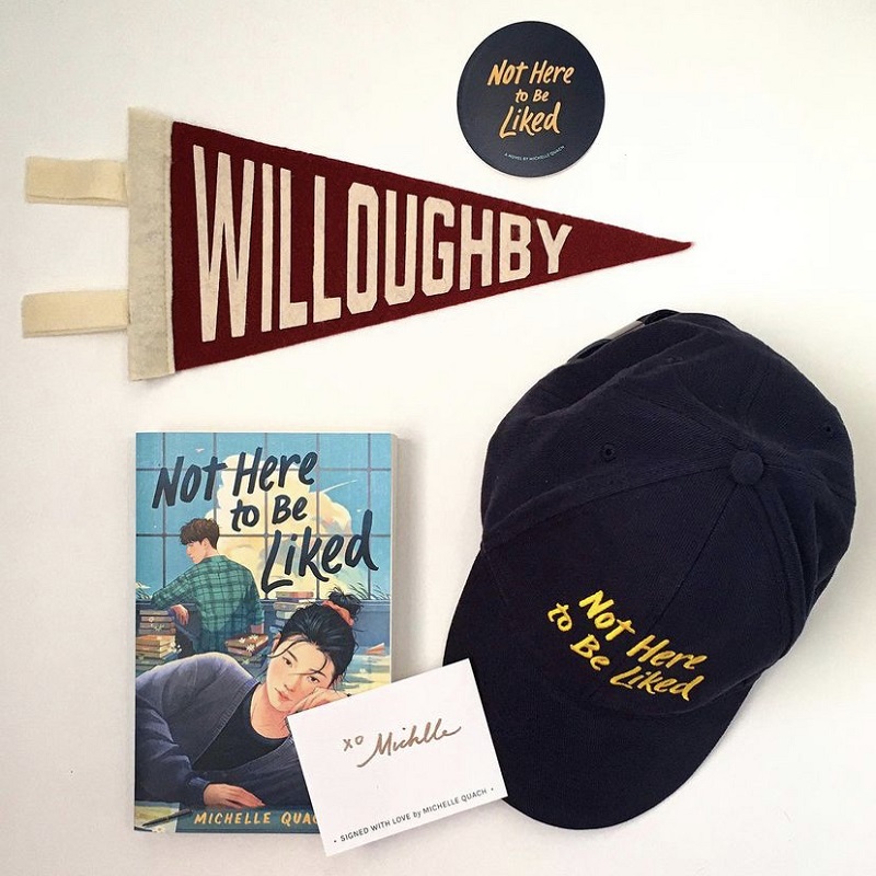 A copy of Not Here to Be Liked with a signed book plate, a black hat embroidered with Not Here to Be Liked, a Willoughby High School pennant, and a round black sticker that says Not Here to be Liked.