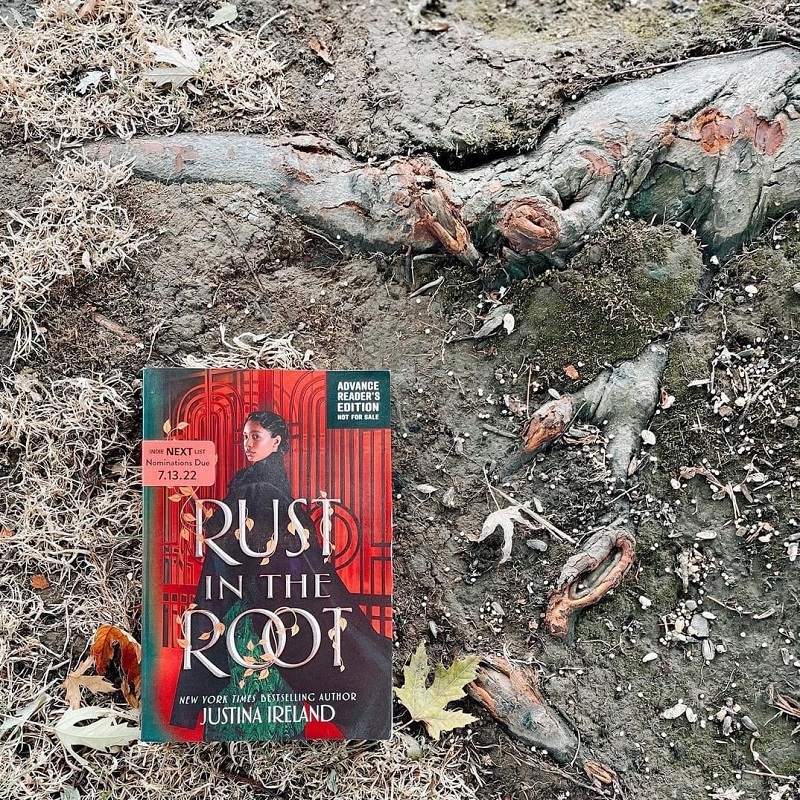 A copy of Rust in the Root lies on the ground surrounded by dead leaves and a large tree root. The cover shows a black girl in a large black robe against a red, industrial-looking gate. The title is scrawled in gold across the cover, and the author’s name sits at the bottom