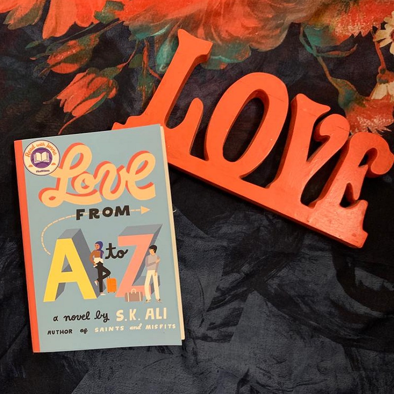 A picture of a paperback copy of Love From A To Z with one corner on top of red, carved wooden letters spelling out “LOVE” laying on a floral hijab.