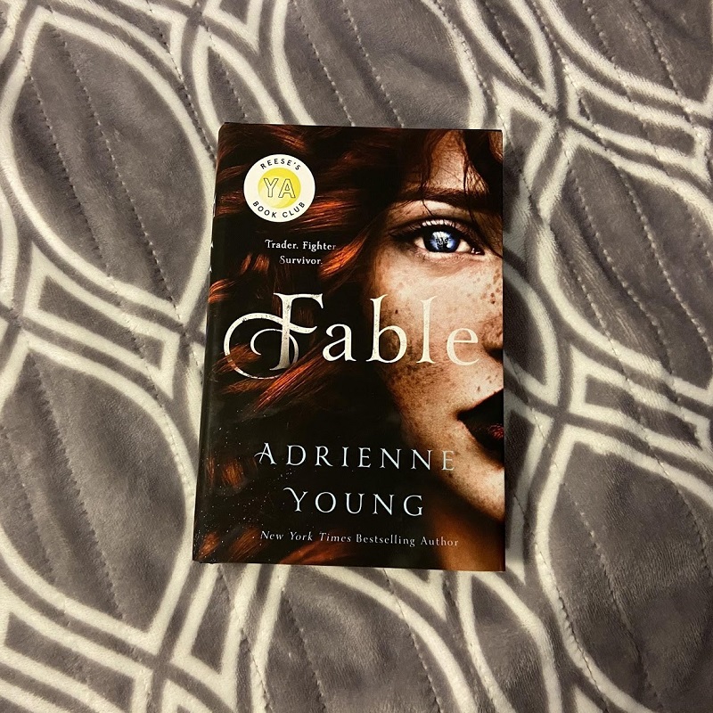 A copy of Fable by Adrienne Young lays face up on a grey and white blanket.