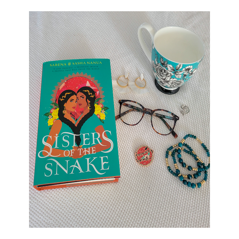 A copy of Sisters of the Snake on a white background with a pair of white fringe earrings, a teal mug with black and white roses, a pair of teal and brown tortoise glasses, an enamel pin, and a stack of teal and gold bracelets.