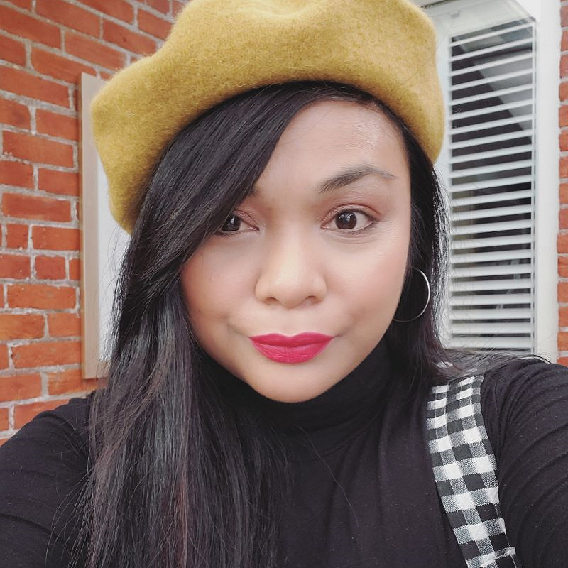Vanessa is wearing a black, long-sleeve turtleneck under a black and white checkered tank top. She has a silver hoop earring visible in her left ear. Her long brown hair is over her right shoulder. She has a yellow beret on her head and is wearing red lipstick. Vanessa is standing in front of a red brick building, with a white windowpane visible.