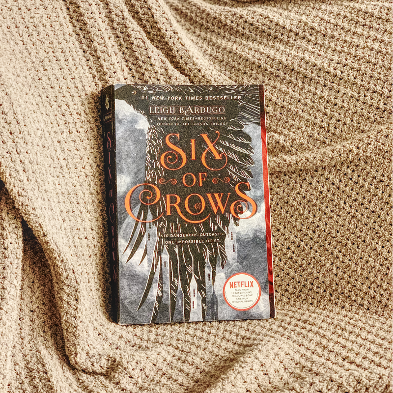 Paperback copy of Six Of Crows against a beige blanket.