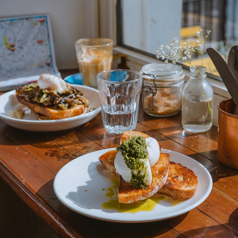 : two plates of eggs on toast sit in the sun at a diner. A water glass sits in between the two plates and a small jar with baby’s breath sits as decoration.