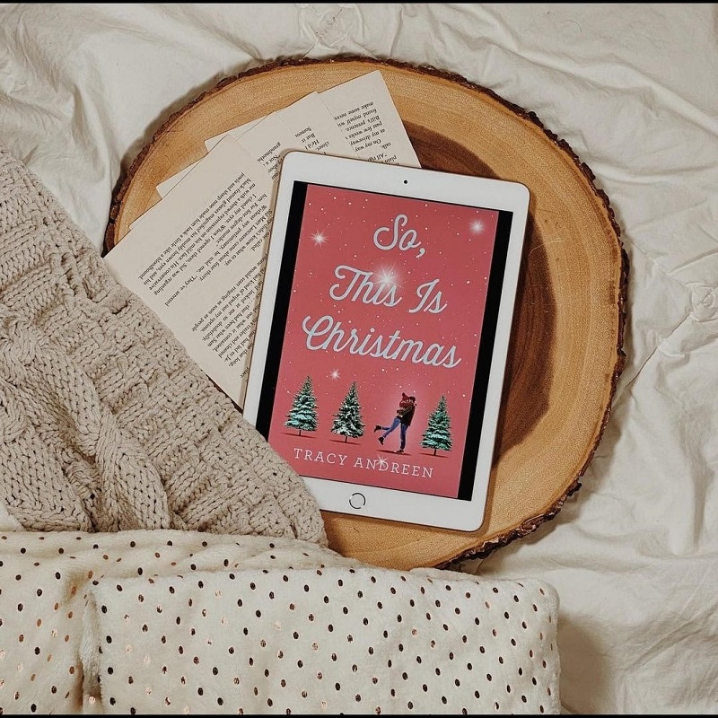 The cover of So, This Is Christmas on an iPad laying on a block of wood on a bed with cozy blankets.